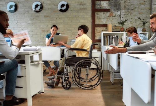 Portrait of young male worker in wheelchair talking to female colleague while presenting ideas using laptop in co-working space. Creative team at work. Disability and handicap concept. Horizontal shot