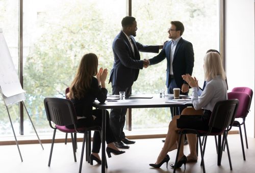 Black chief ceo shaking hand of millenial european worker manager welcoming or thanking for good job while staff applaude, happy young businessman is awarded by boss in presence of colleagues